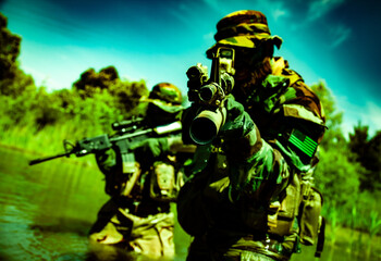 Two soldiers in camouflage uniforms and helmets aim rifles in a lush, green landscape, partially submerged in water with trees in the background.