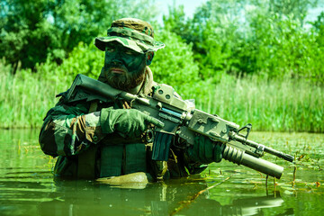 Soldier in camouflage holding a rifle partially submerged in water with green vegetation in the...