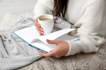 Young woman with blanket and cup of coffee reading book at home, closeup