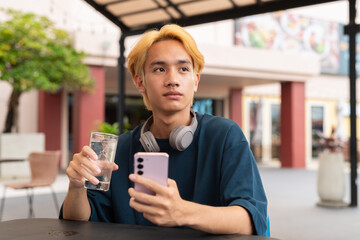 Asian gay teenager drinking water and using phone outdoors in cafe restaurant