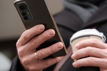 Handsome man wearing black coat and brown leather bag walking down on the city, drinking coffee paper takeaway cup with mobile phone. Stylish bearded middle-aged male using smartphone at urban street
