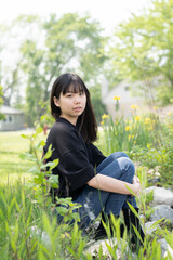 Young asian teenage girl sitting on rock in a grassy area, surrounded by green plants and wildflowers, wearing a black top and jeans.