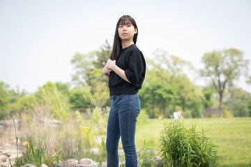 Young asian teenage girl standing on the ground in a grassy area, surrounded by green plants and wildflowers, wearing a black top and jeans.