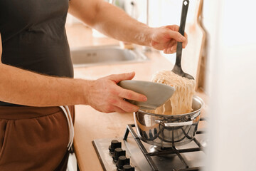 Man cooking ramen noodles. Male making noodle soup broth in the home kitchen stove. Traditional Asian food. Close Up Photo Of Mans Hands Serving Pasta With Fresh Vegetables. Preparing Homemade Dish.