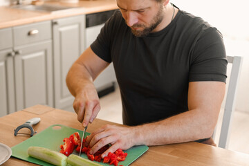 Man preparing fresh vegetables in modern kitchen, organic ingredients. Healthy meal. Cooking Food At Home. Male slicing red pepper and green cucumber on a wooden cutting board. Nutritionist, culinary