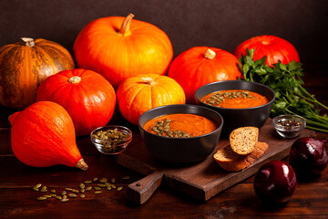 Delicious pumpkin soup garnished with seeds, served with bread on a wooden board, surrounded by vibrant pumpkins, red onions, and parsley. Cozy and rich autumnal culinary setting.