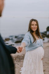 Happy pregnant woman holding partner’s hand on beach, smiling at camera. Maternity love, outdoor lifestyle and family expecting baby