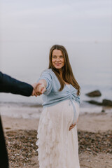 Happy pregnant woman holding partner’s hand on beach, smiling at camera. Maternity love, outdoor lifestyle and family expecting baby