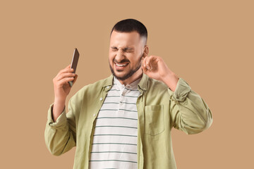 Young man with mobile phone suffering from loud noise on beige background