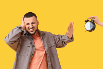 Young man suffering from loud noise and hand with alarm clock on yellow background