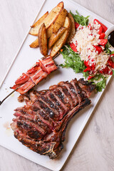 Bistecca di carne cotta alla brace con cottura al sangue accompagnata da uno spiedino di maiale  e con contorno di patate al forno, rucola, formaggio e pomodori servita in una braceria