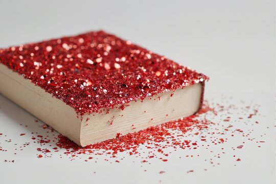 Close-up of a closed white book with its cover covered in red glitter in the shape of hearts. On a white background