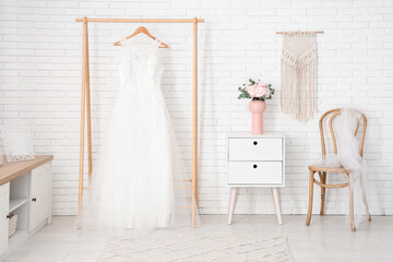 Vase with bouquet of beautiful ranunculus flowers near wedding dress on hanger at home