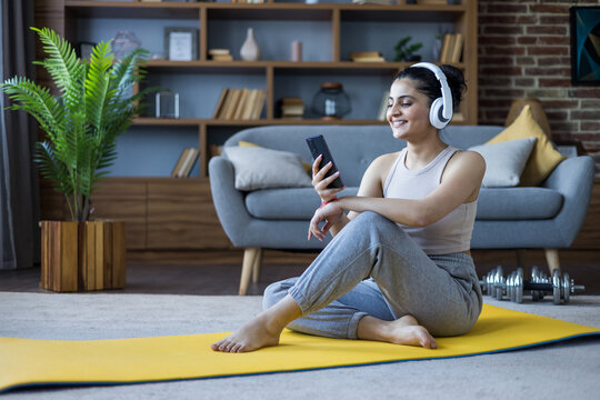 A young Indian girl is exercising at home, sitting on a mat, wearing headphones, and looking at a mobile phone in her hand