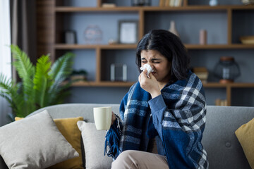 An Indian young woman is sick and sitting at home on the couch covered with a blanket, wiping her...