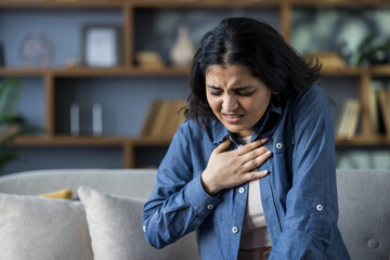 A young Indian woman sits on a couch at home, holding her hand over her heart, grimacing as she feels severe pain in her chest