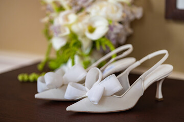 A pair of elegant white bridal heels with satin bows rest gracefully on a wooden surface, with a...