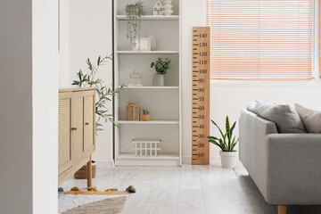 Interior of modern living room with sofa, shelving unit, commode and wooden stadiometer near window
