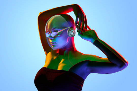 Portrait of bald woman wearing 3D futuristic clear helmet and goggles, arms raised in expressive gesture under stylized neon lighting and clear blue backdrop. Cyberpunk cover art, social media