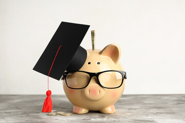 Piggy bank in eyeglasses with graduation hat and money on grunge grey table near wall. Tuition fees...