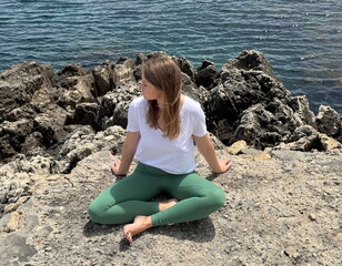 A woman sits peacefully on coastal rocks, soaking in the ocean breeze and practicing mindful...