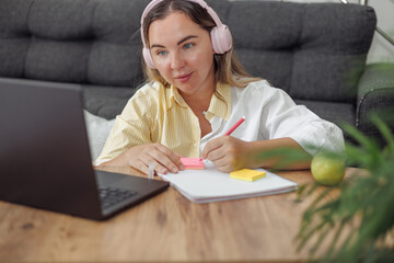 Young blond European woman with headphones sitting with laptop and notebook, studying or working...