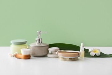 Composition with cosmetic products, massage brushes and plant leaf on light table against color wall