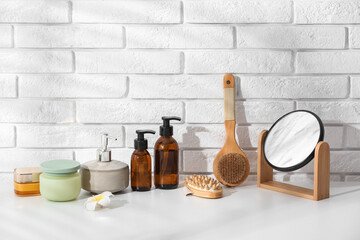 Set of different cosmetic products, mirror and massage brushes on table near light brick wall