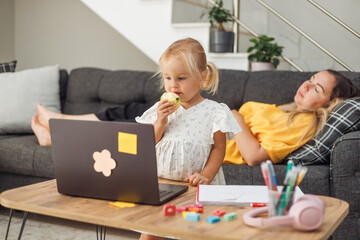 Young blond European girl eating an apple near a laptop on a table. Mother woman with dark hair...