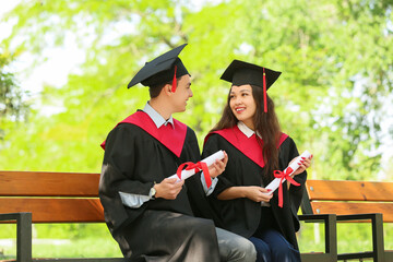 Couple of graduating students with diplomas sitting on bench outdoors