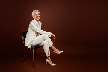 Confident mature woman in stylish formal outfit sitting on a chair against a brown background...