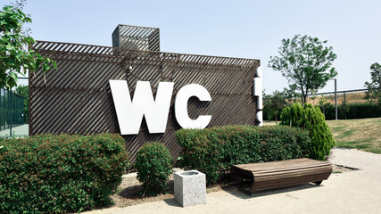 Fototapeta premium Modern public toilet building with large WC letters on facade, surrounded by greenery and bench, concept of urban infrastructure, theme of public amenities, city life