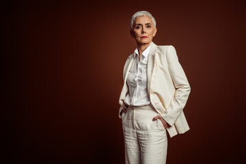 Confident mature businesswoman in elegant suit with white shirt posing against a brown background, exuding professionalism