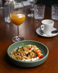 A bowl of yogurt topped with granola, fruits, and nuts alongside a glass of juice on a wooden table during breakfast.