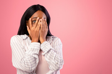 Portrait of young black woman covering face with hands, feeling afraid or stressed on pink studio...