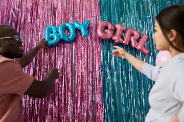 Young Black man and young Asian woman arranging metallic boy and girl balloons against pink and...