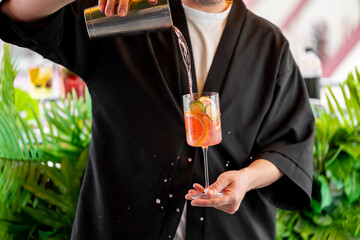 A bartender pours a refreshing cocktail into a glass with citrus slices and cucumber. The vibrant...