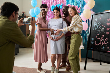 Friends smiling and touching pregnant woman belly while man photographing them with smartphone at...