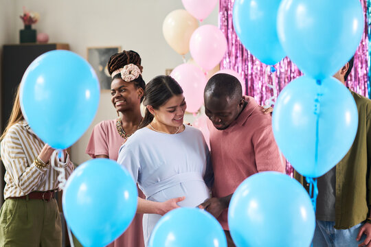 Young adult Caucasian woman with pregnant belly standing beside Black man gently touching her stomach, surrounded by multiethnic friends - Powered by Adobe