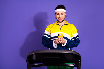 Young man wearing vibrant sportswear looking at his smartphone while standing on a treadmill in front of a purple background