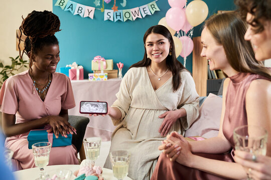 Pregnant young adult Caucasian woman sitting with diverse group of young adult women showing ultrasound image on smartphone