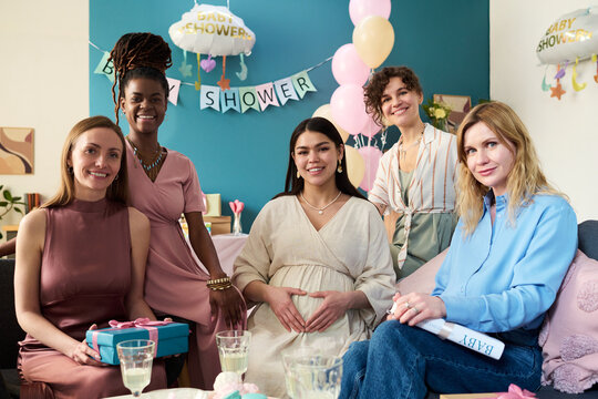 Group of young adult women including Caucasian and Black women smiling and posing together at gender reveal party with pregnant woman sitting in center - Powered by Adobe