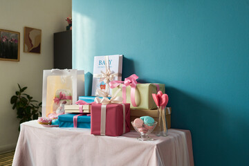 Gift boxes with pastel ribbons, baby-themed book, sweets and decorative items arranged on table against blue wall, celebrating gender reveal party with festive presentation