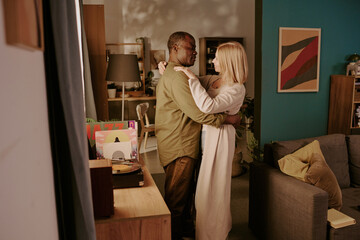 Senior Black man and senior Caucasian woman embracing in living room, standing close together and looking at each other, expressing romance and affection in home setting