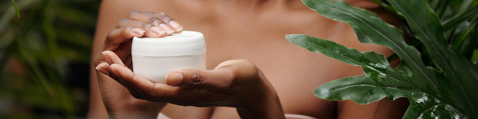 Close-up of hands of unknown girl holding white jar of moisturizing cream surrounded by lush green leaves. Woman preparing to use skincare product in natural setting