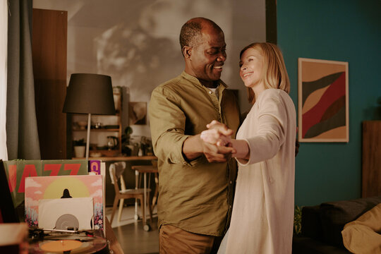 Senior Black man and senior Caucasian woman dancing together in living room, smiling and holding hands, enjoying romantic moment, standing close and looking at each other