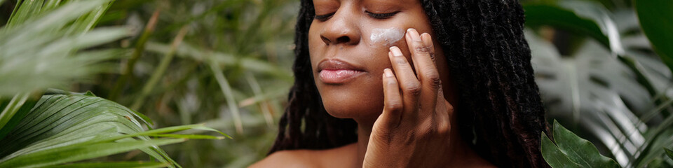 Woman closing eyes while gently applying skincare product surrounded by lush green plants creating...
