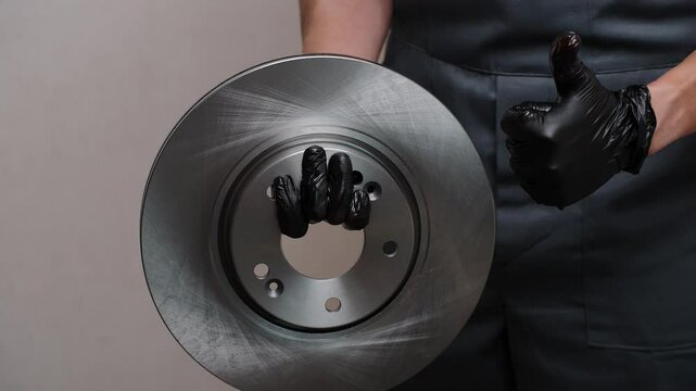 Auto mechanic in black gloves holds new brake disc in hands, runs fingers over it and shows thumb up, close-up. Auto parts replacement concept, car maintenance, quality parts, car brake system.