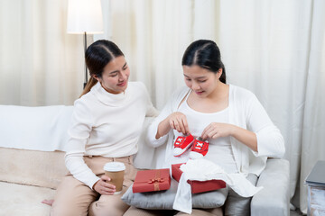 Asian pregnant woman happily opening baby gift box from friend sitting together on sofa expressing joy excitement appreciation celebrating baby’s arrival wearing casual white sweater brown pants