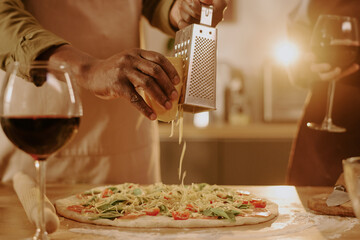 Senior Black man grating cheese onto homemade pizza while senior Caucasian woman holding glass of red wine, both preparing meal together in kitchen, hands visible, romantic atmosphere implied